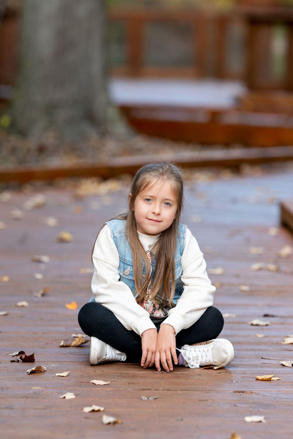 Young girl with long hair sits cross-legged on a wooden boardwalk covered in fall leaves.