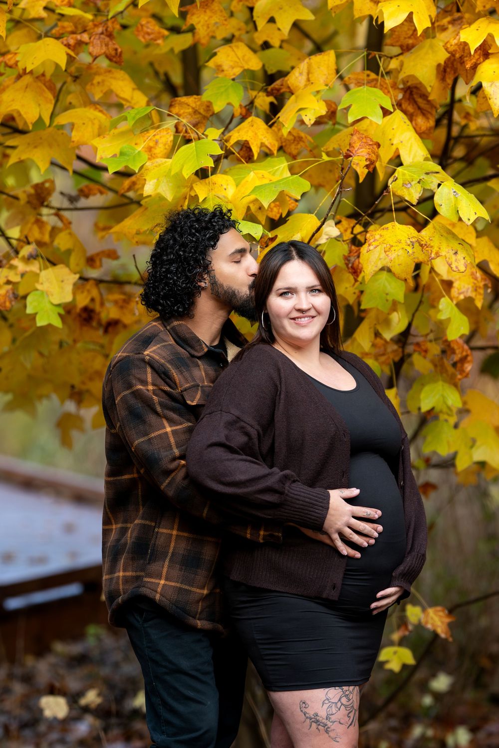 Man kisses pregnant womans cheek as they embrace under bright yellow fall maple tree.