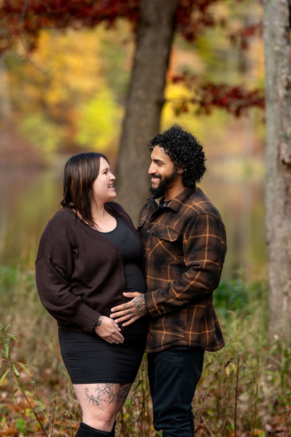 Pregnant couple shares a loving gaze while cradling baby bump amid vibrant autumn trees by a lake.