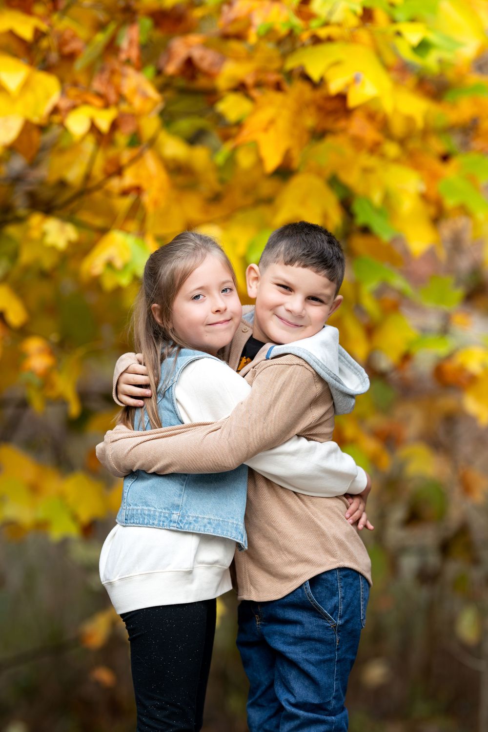 Two children hugging and smiling in front of vibrant yellow autumn foliage outdoors.