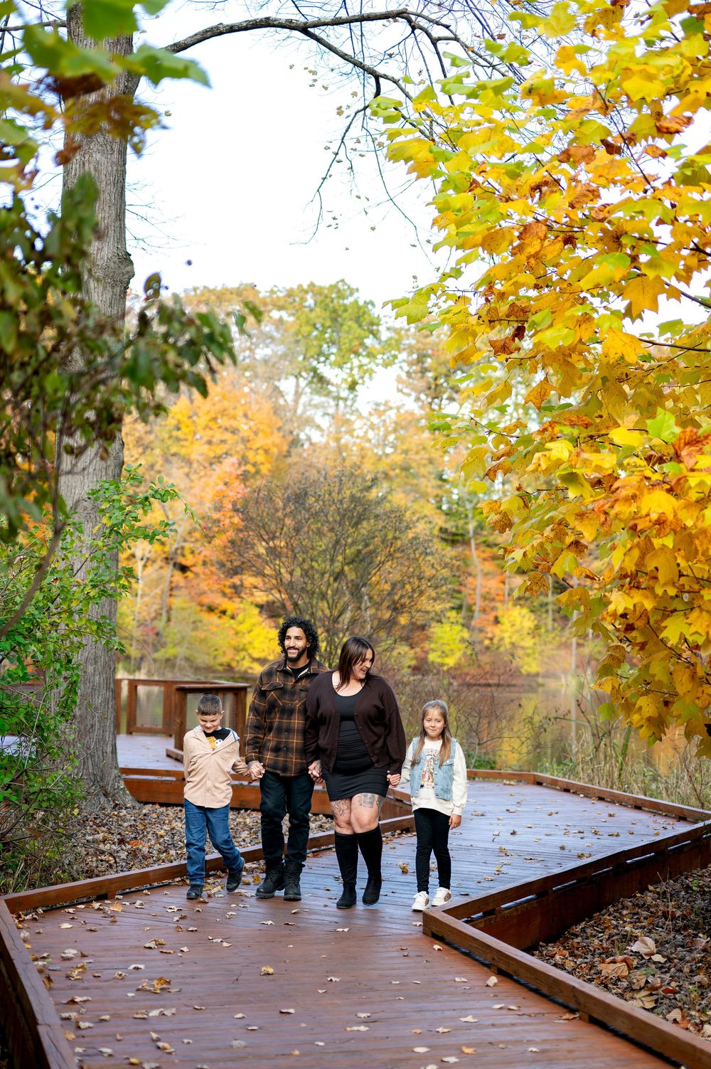 Family of four walking together on a wooden boardwalk amid bright autumn trees.