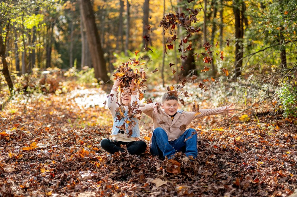 Two children joyfully tossing autumn leaves while sitting on the ground in a forest.