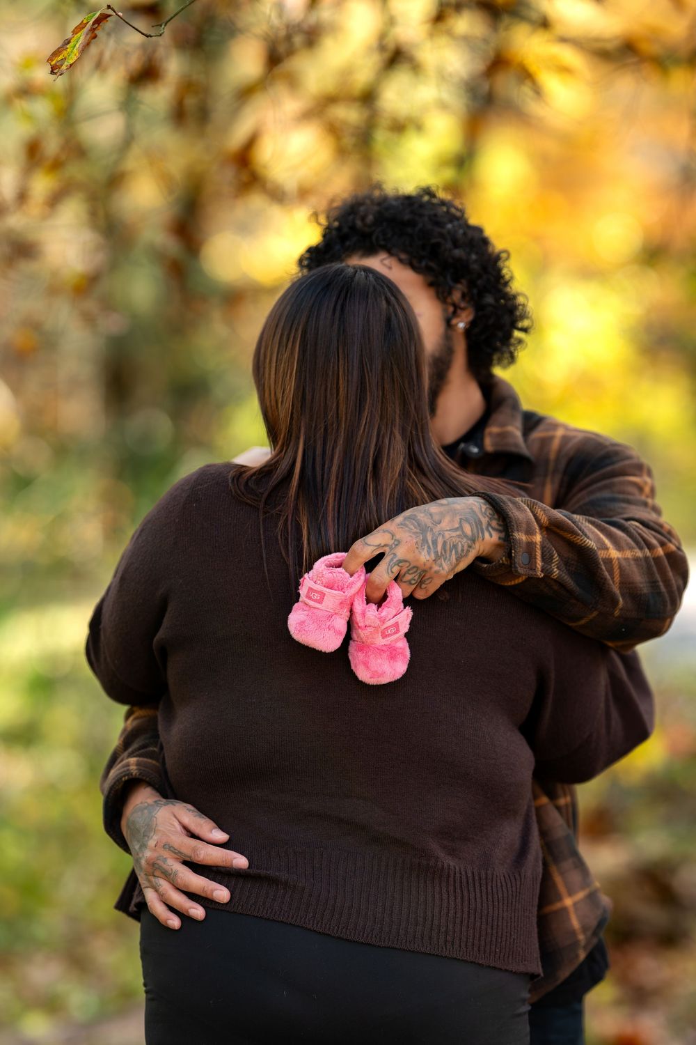 Couple embracing outdoors holding pink baby booties against golden autumn foliage backdrop.