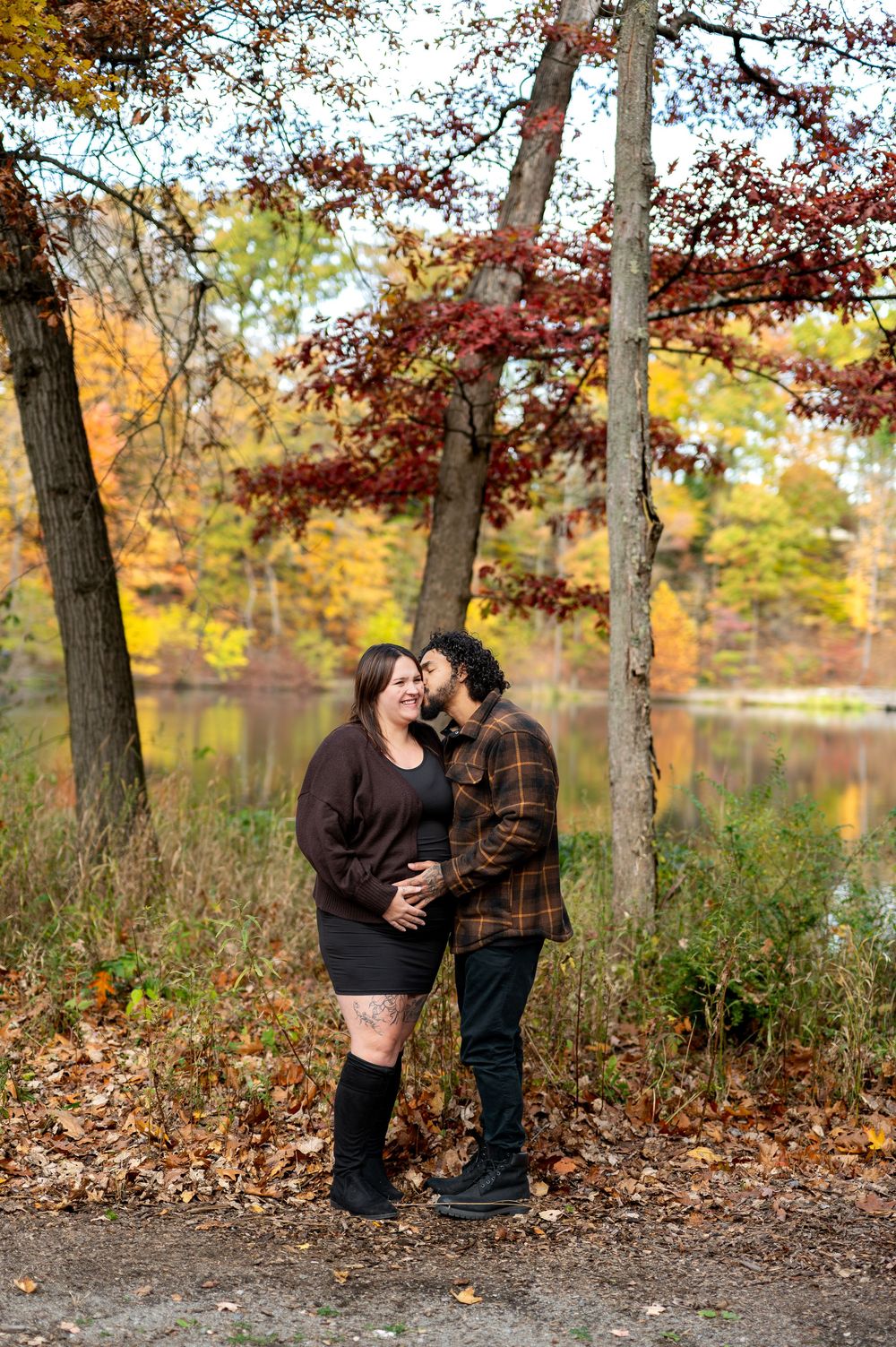 Man tenderly kisses pregnant woman's cheek as they stand among colorful fall foliage near a lake.