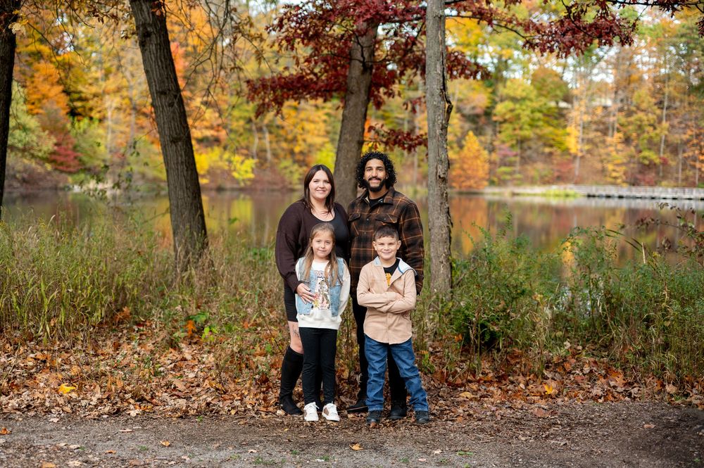 Family of four stands together by a lake surrounded by brilliant autumn foliage in warm earth tones.