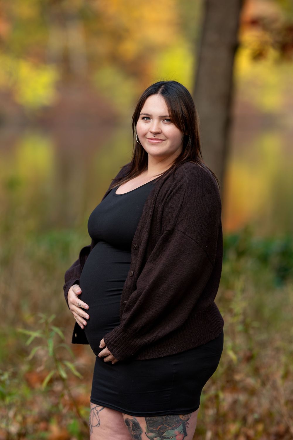 Pregnant woman in black dress and brown cardigan cradles baby bump in a colorful autumn forest setting.
