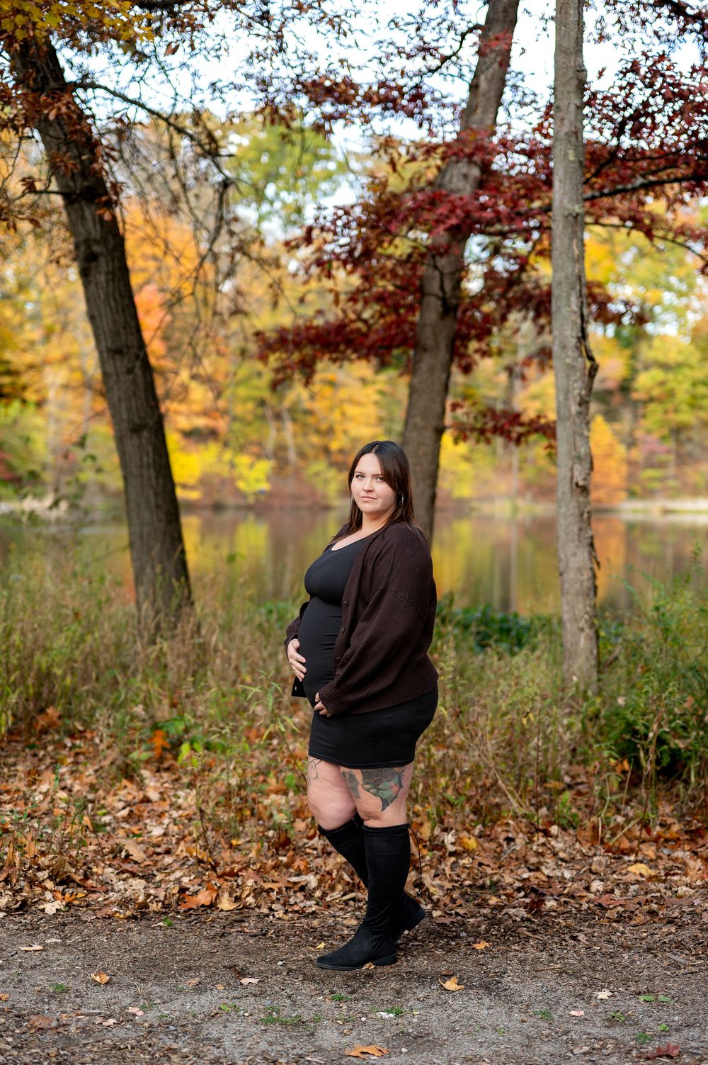 Pregnant woman in brown cardigan and knee-high boots poses in autumn park by a lake.