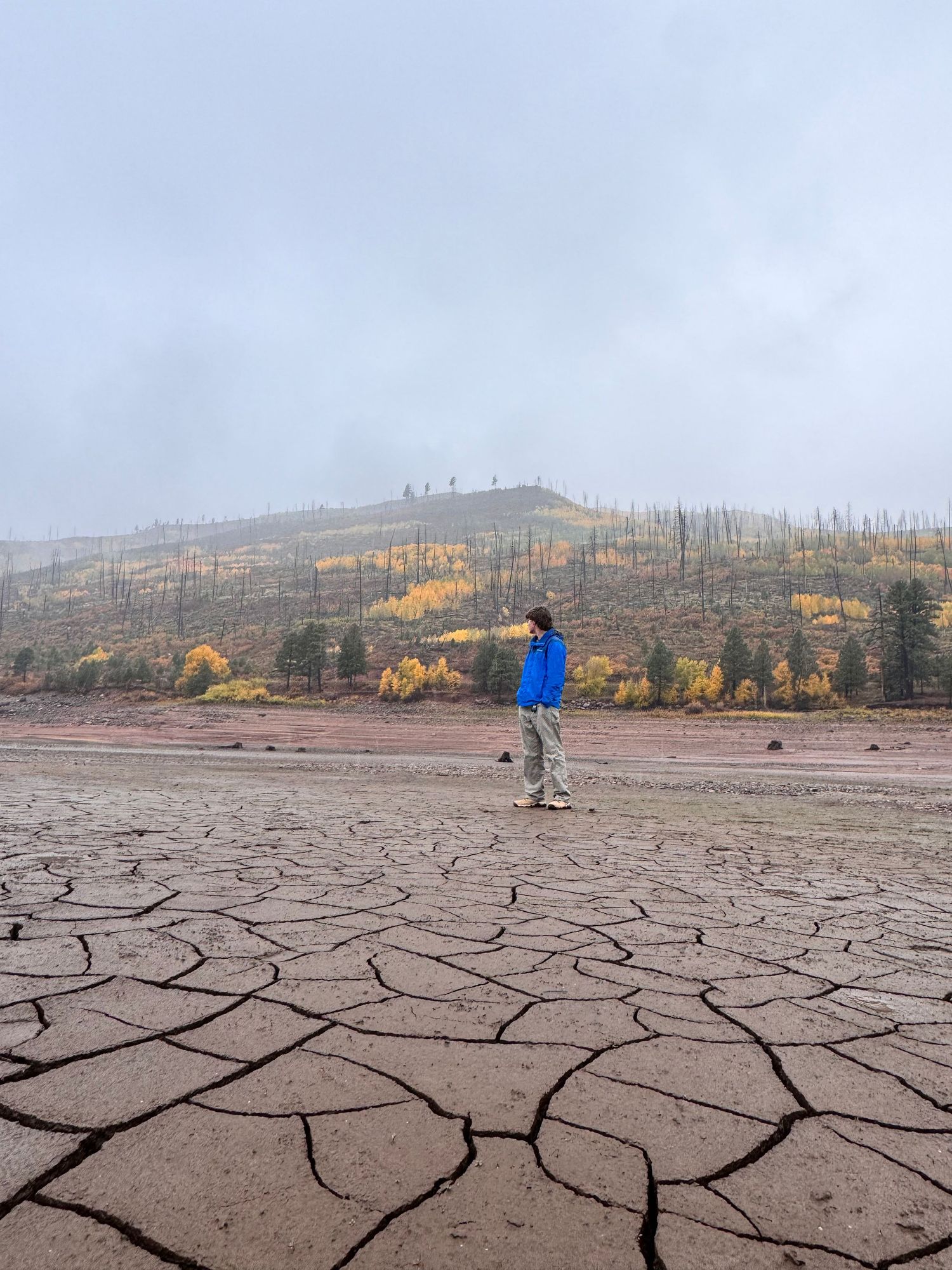 A cracked and dry lakebed stretches across the landscape with autumn trees and hills visible in the background.