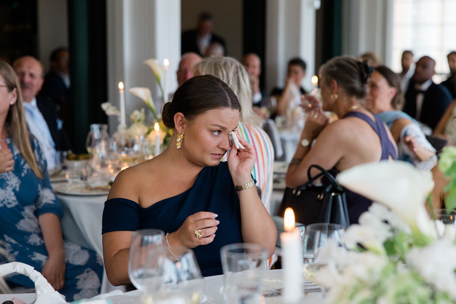 Wedding reception guests seated at elegant tables with white linens and floral centerpieces enjoy dinner and conversation.