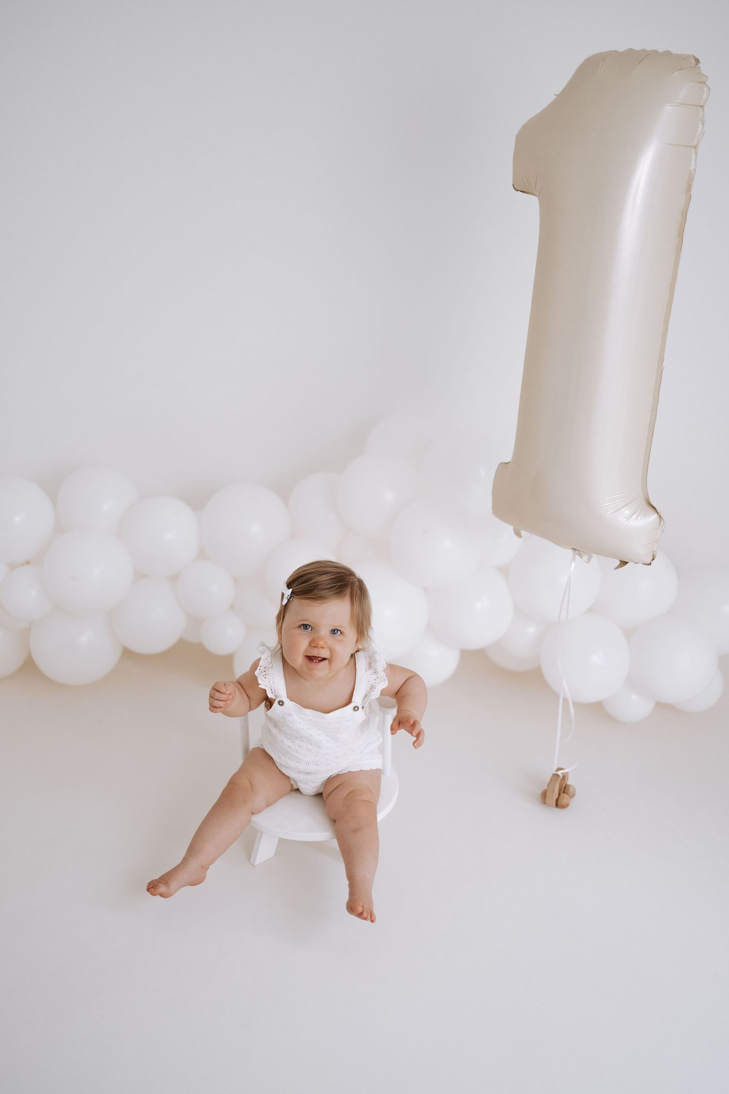 Four minimalist white photos showing a baby in different poses against a white backdrop with white balloon decorations.