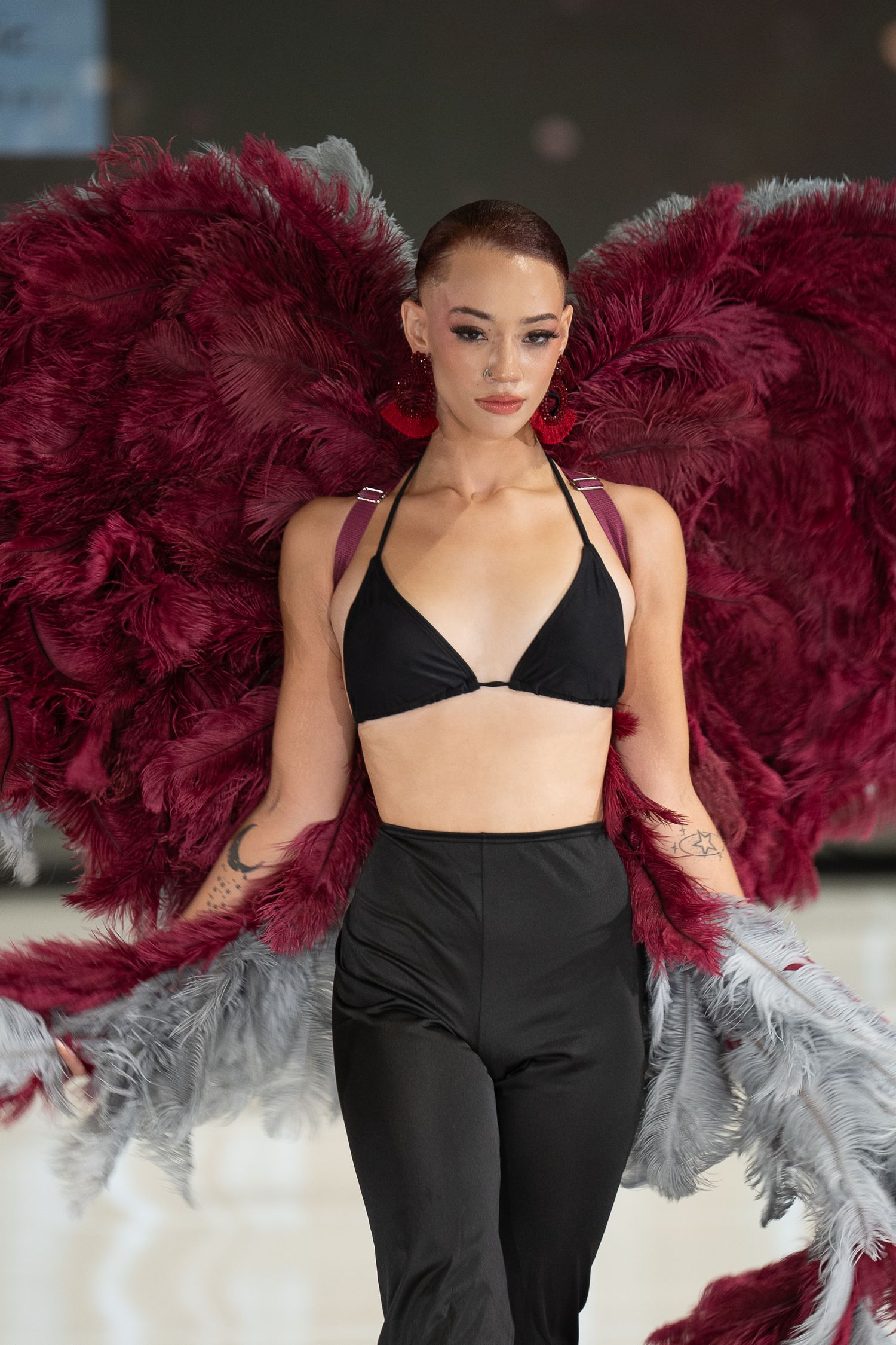 Fashion model poses in black outfit against dramatic burgundy and white feathered backdrop on runway.