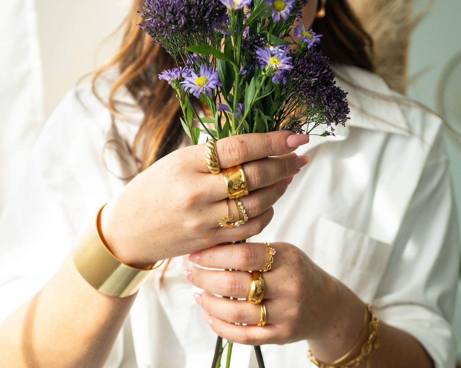 Close-up of woman in white shirt holding purple flowers, wearing multiple gold rings and bracelets.