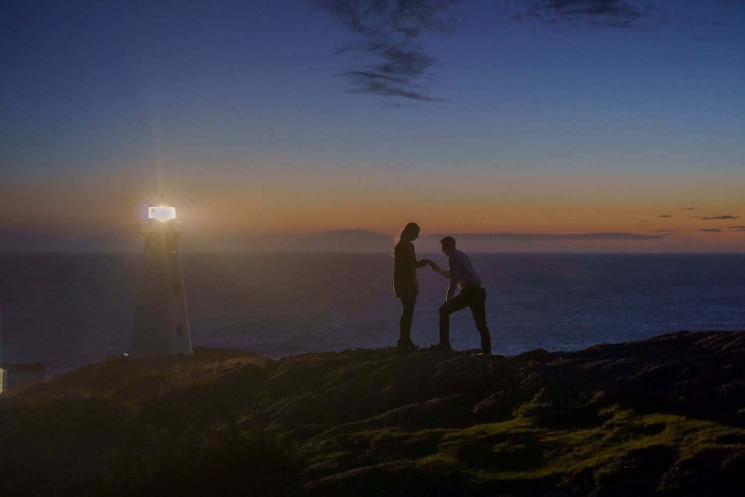 Proposal photo at the Cape Spear lighthouse with the rising sun over the ocean, orange dawn light, and a cold blue morning sky.