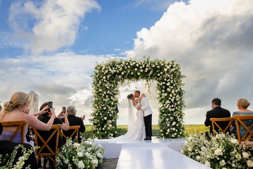 A romantic outdoor wedding ceremony under a floral arch with guests seated on white chairs in a garden setting.