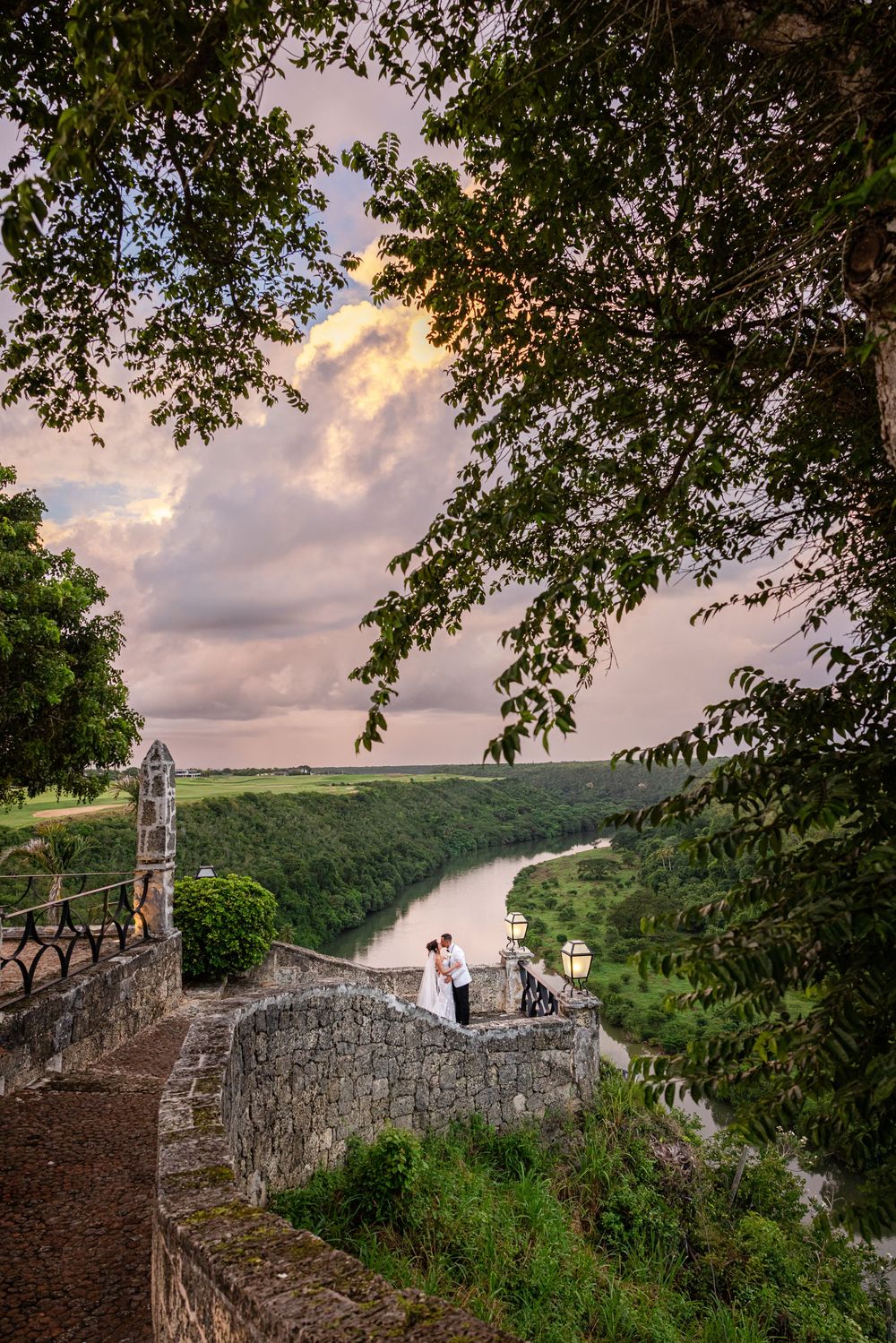A scenic overlook at sunset shows a stone wall path winding along a river valley with lush green forest views.