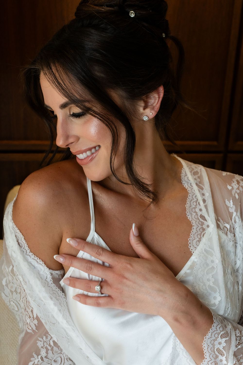A bride in white lace smiles joyfully while wearing a delicate robe and pearl earrings.