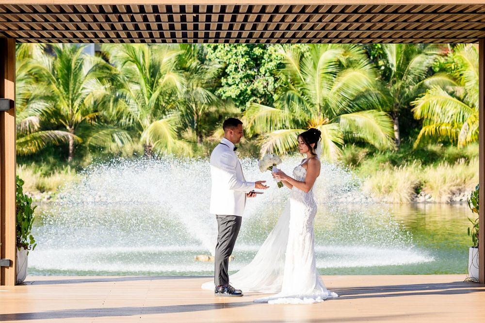 Couple in formal attire sharing a romantic moment under a pergola with tropical palm trees and fountain in background.