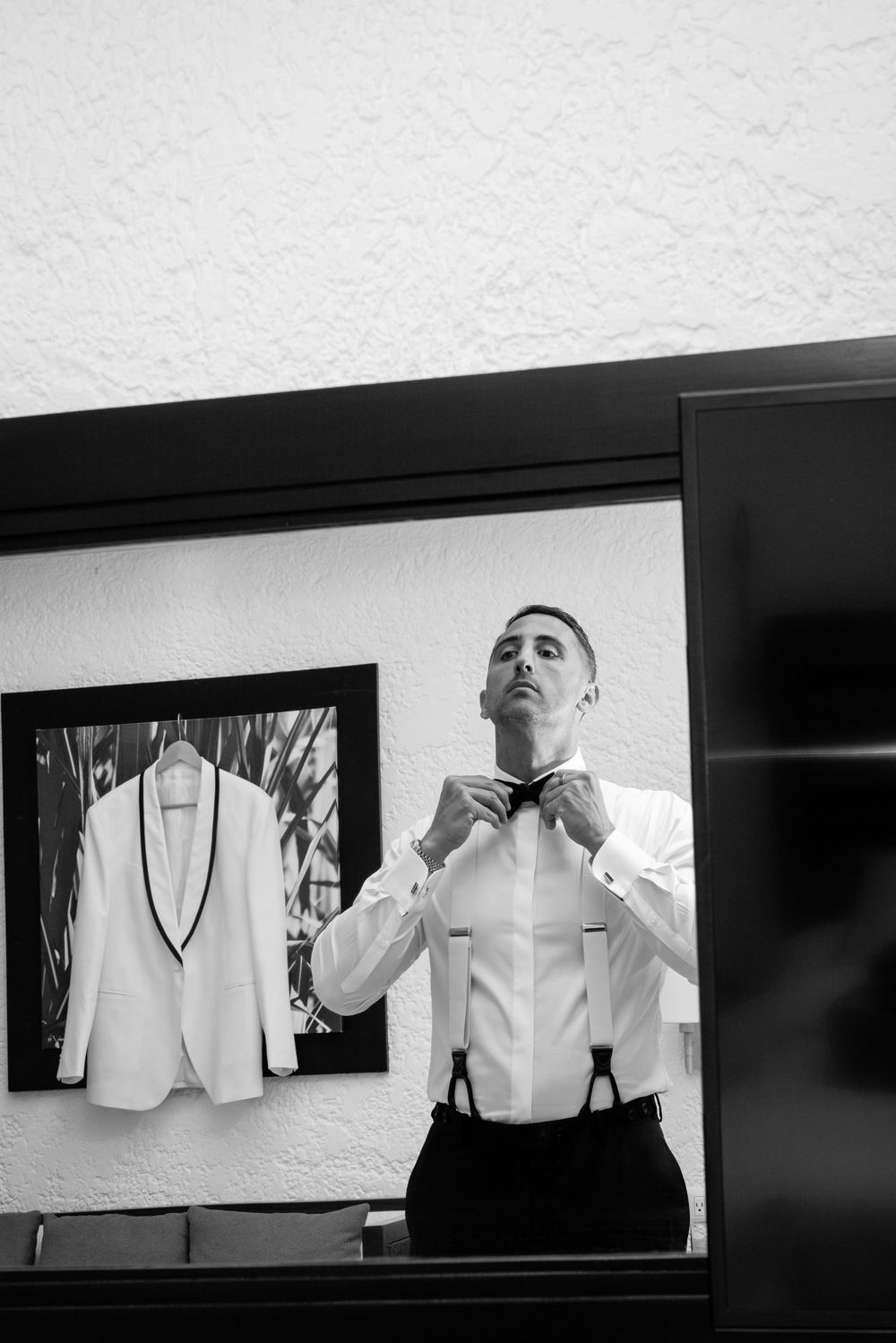 Black and white photo of groom adjusting bow tie in mirror with white tuxedo jacket hanging in background.
