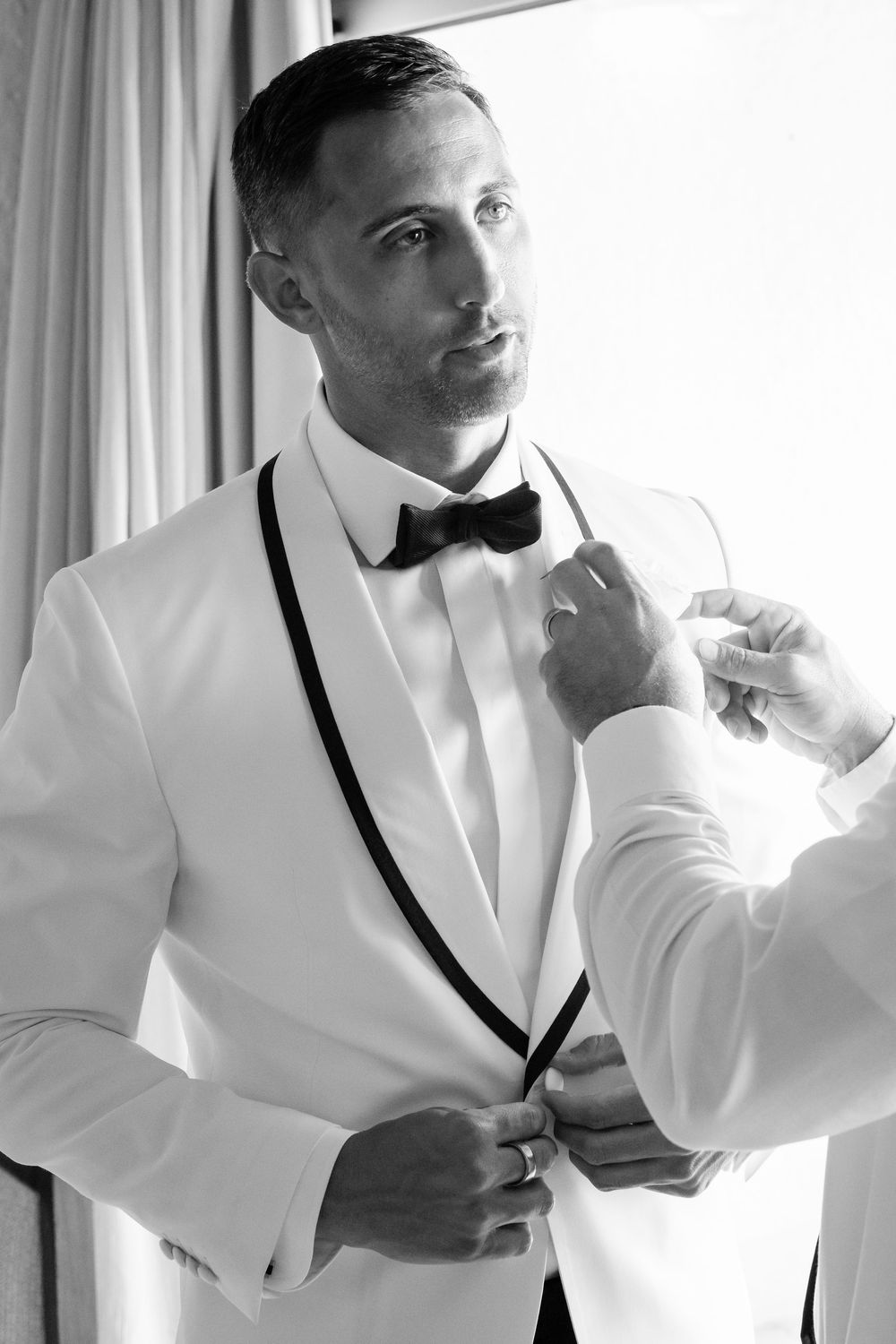 Groom in white tuxedo with black bow tie being helped to dress in black and white photo.