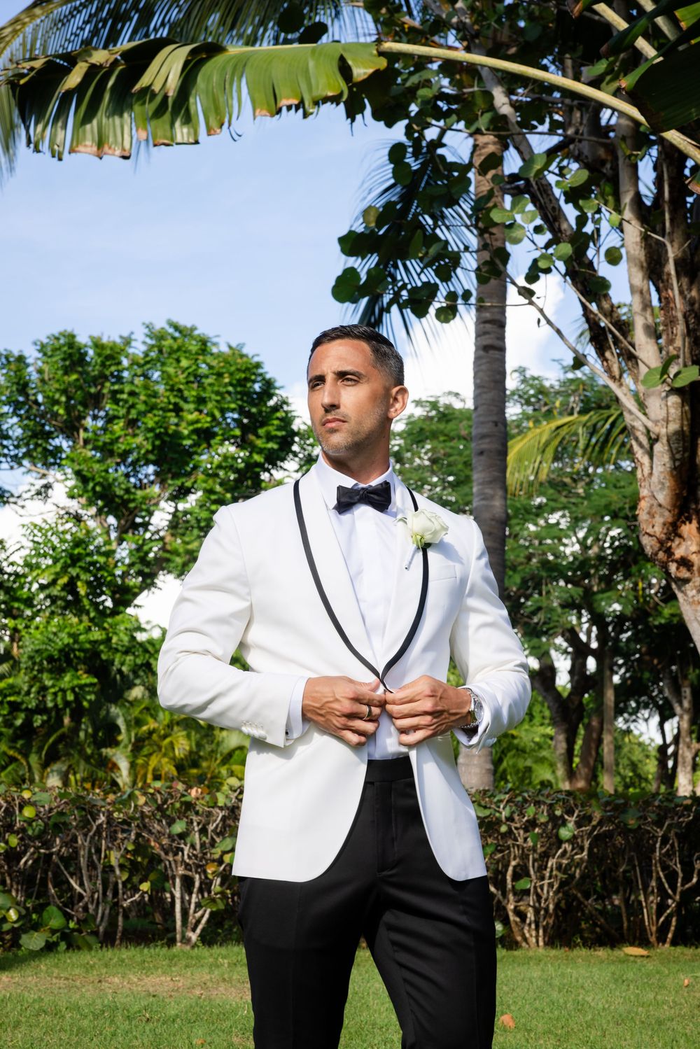 Groom in white tuxedo jacket with black lapels and bow tie, wearing boutonniere, standing outdoors among tropical palms.