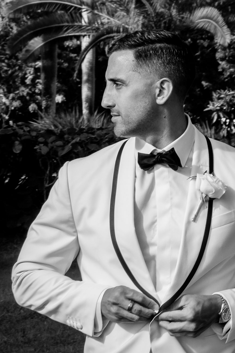 Black and white photo of a groom in a white tuxedo with bow tie adjusting his jacket outdoors.