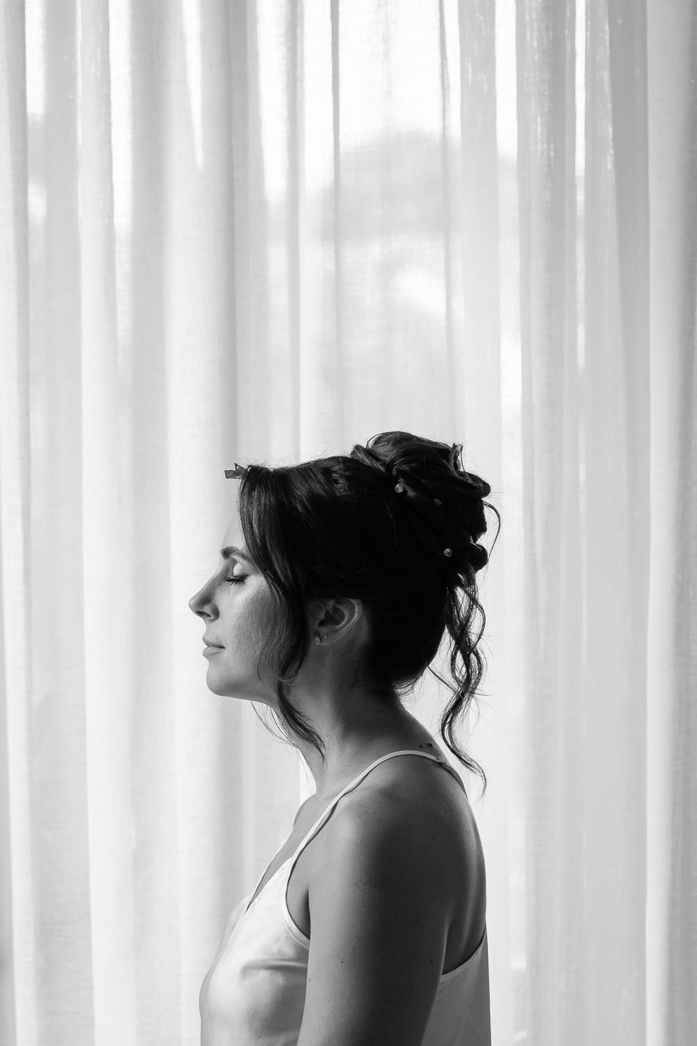 Black and white profile portrait of a bride with elegant updo hairstyle standing before sheer white curtains.