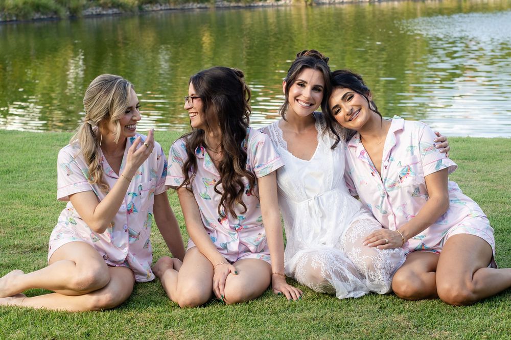Four women in matching floral robes sit on grass by a lake, smiling together in a bridal party photo.