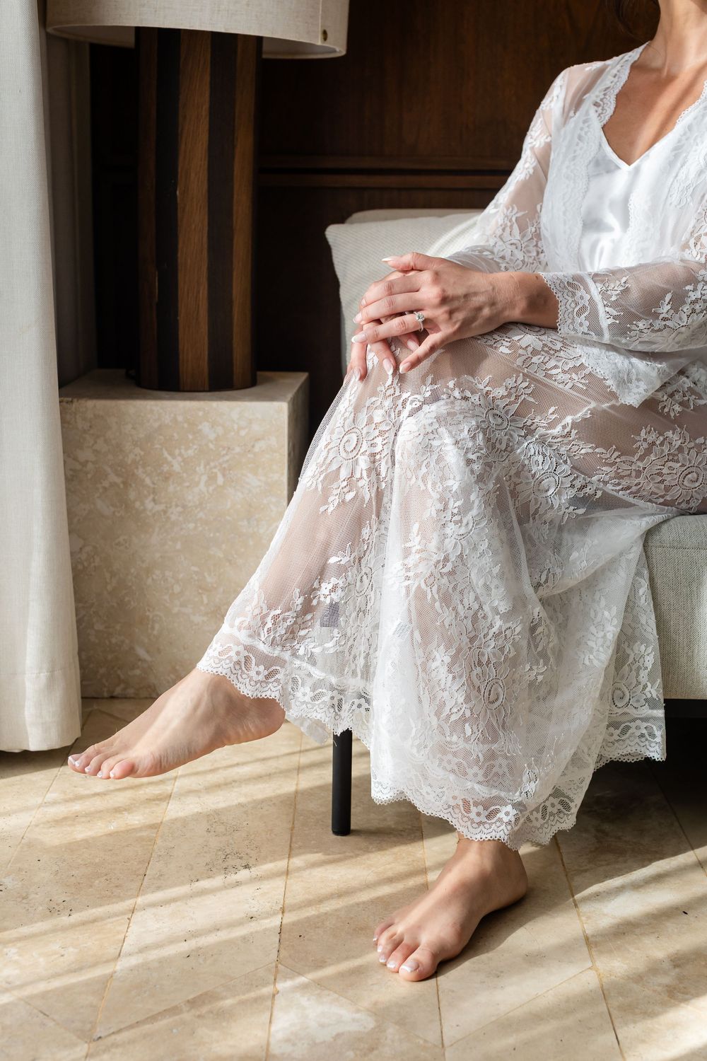 Woman in elegant white lace robe sitting barefoot on a chair, hands folded gracefully in her lap.