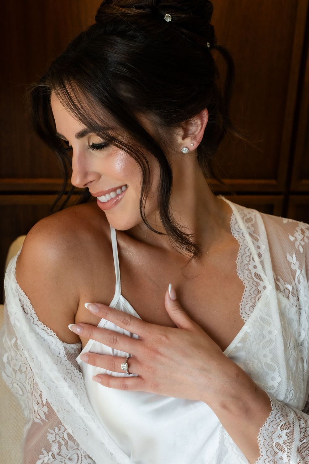 Smiling bride in white lace robe with updo hairstyle and diamond earrings on her wedding day.