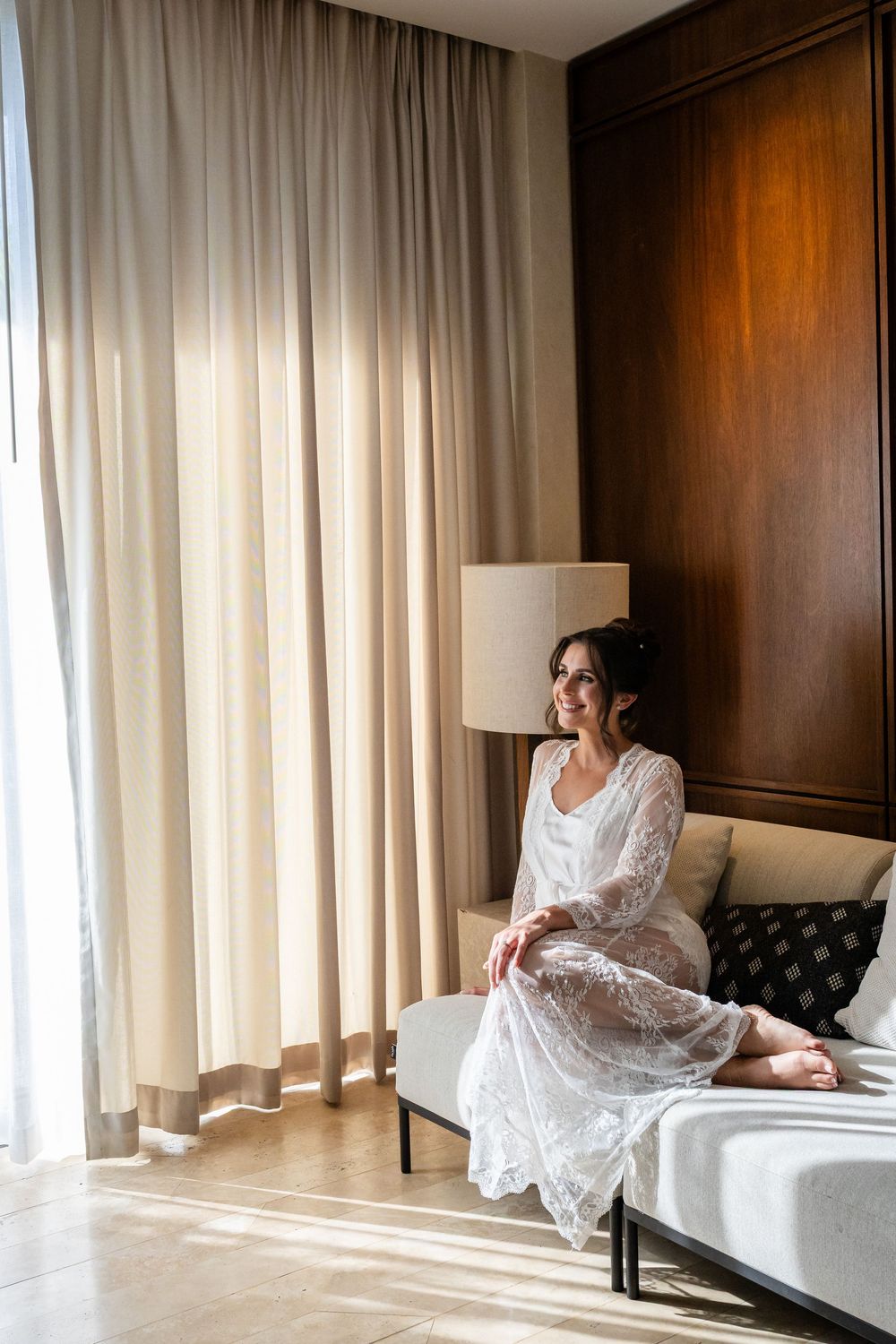 A smiling bride in a white lace robe sits on a bed near sheer curtains in a sunlit hotel room.