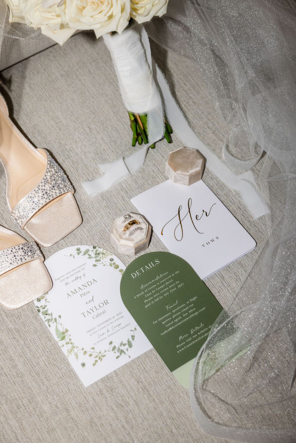 Elegant wedding flat lay with floral invitations, glittery heels, ring box, and white rose bouquet on silver fabric.