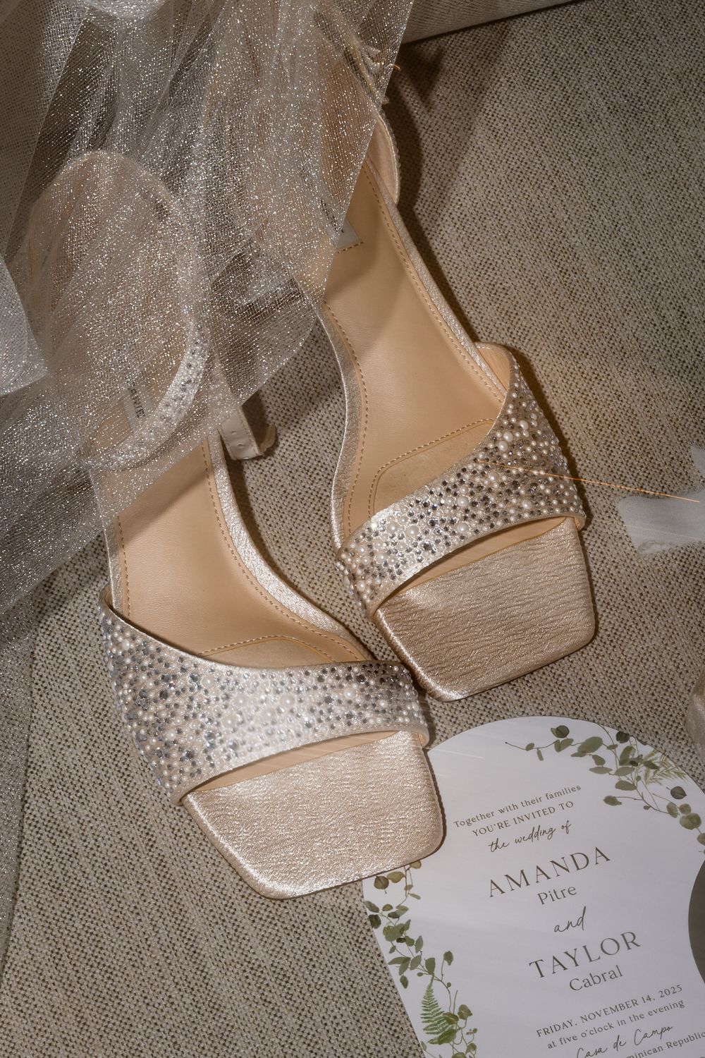 Elegant rhinestone-embellished champagne bridal heels beside a wedding invitation for Amanda and Taylor on a linen surface.