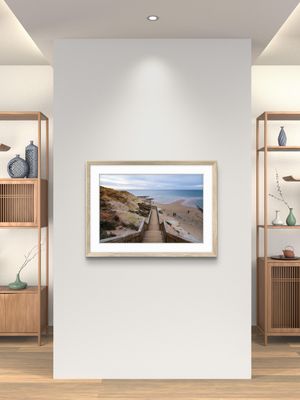 Framed coastal beach path photograph in natural wood frame displayed on white wall in modern minimalist interior.