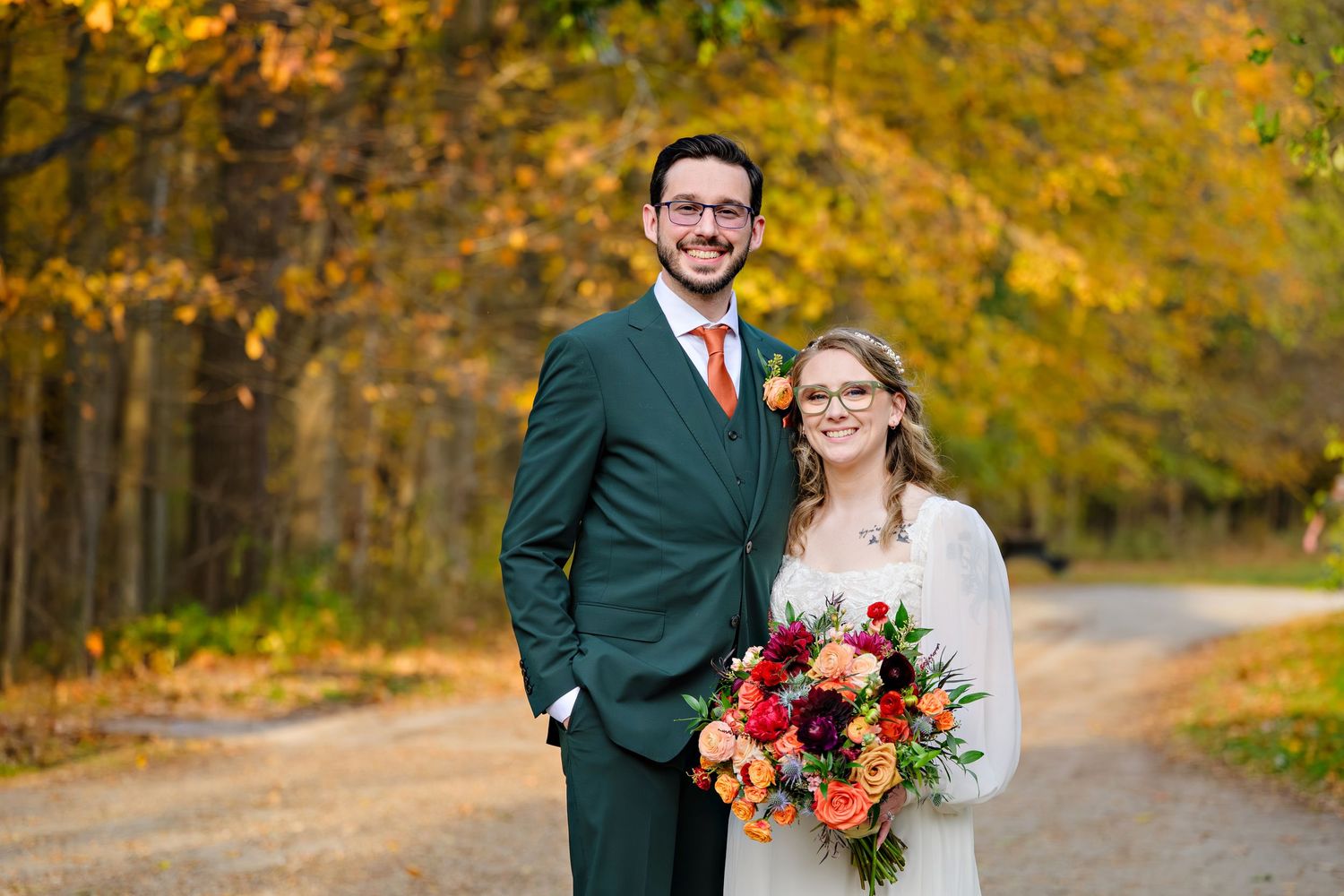 A couple in wedding attire poses together on a forest path surrounded by vibrant autumn foliage at Everett Road Covered Bridge in Ohio.