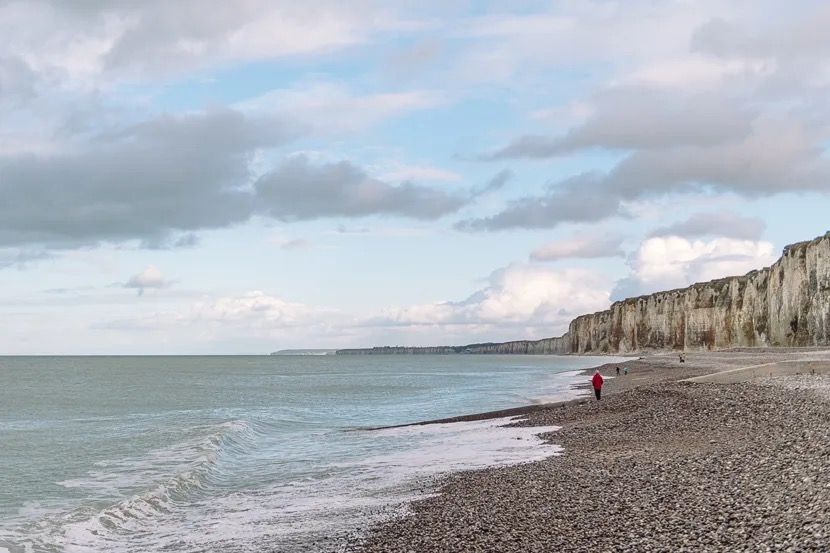 Pebble beach with gentle waves and tall coastal cliffs under a cloudy sky.