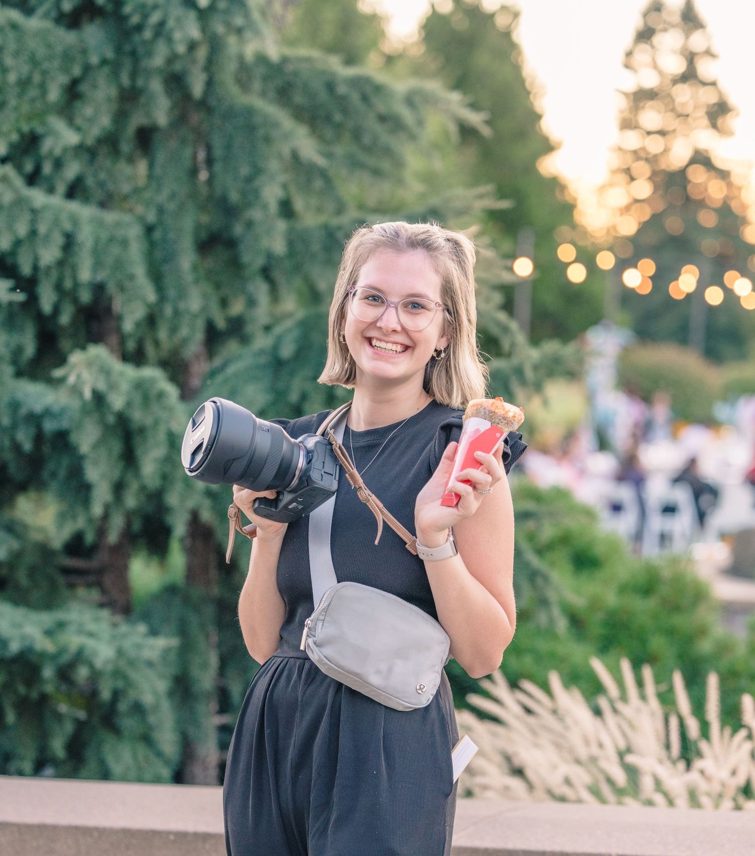 Professional photographer in black holding camera and phone during outdoor photoshoot at sunset.