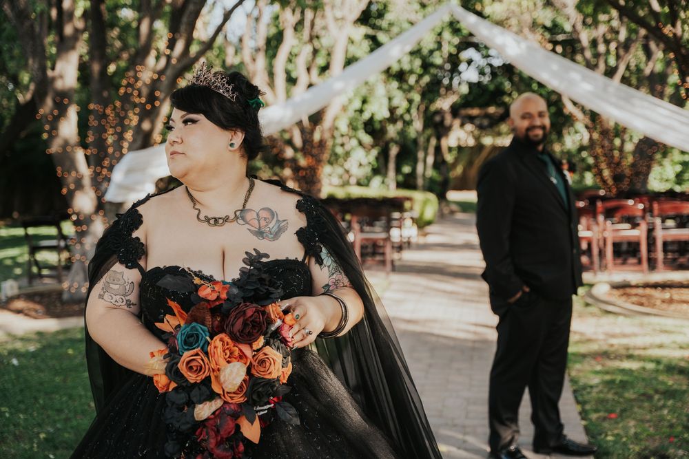 A wedding party poses outdoors in a garden setting with the bride wearing a black dress and holding a colorful bouquet.