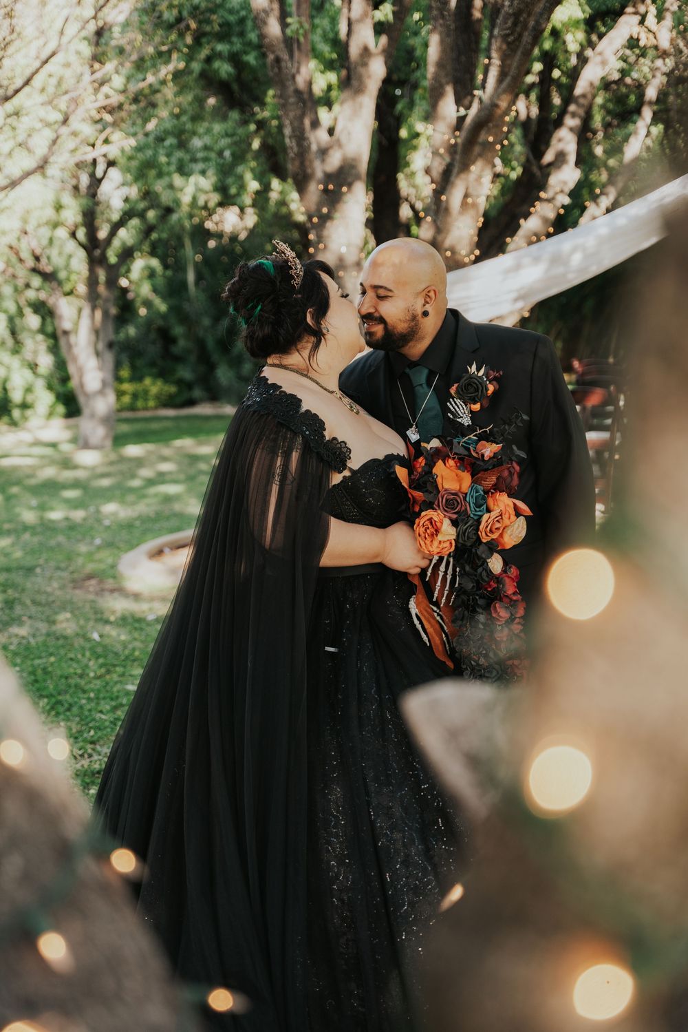 A romantic couple in black attire share intimate moments in a sunlit garden setting with glowing string lights.