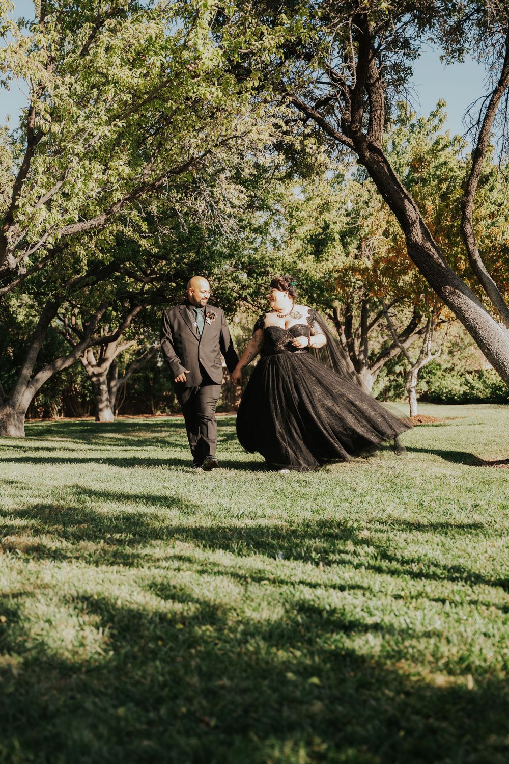 A couple walks together on a sunny day through a peaceful park with twisted olive trees casting dappled shadows.