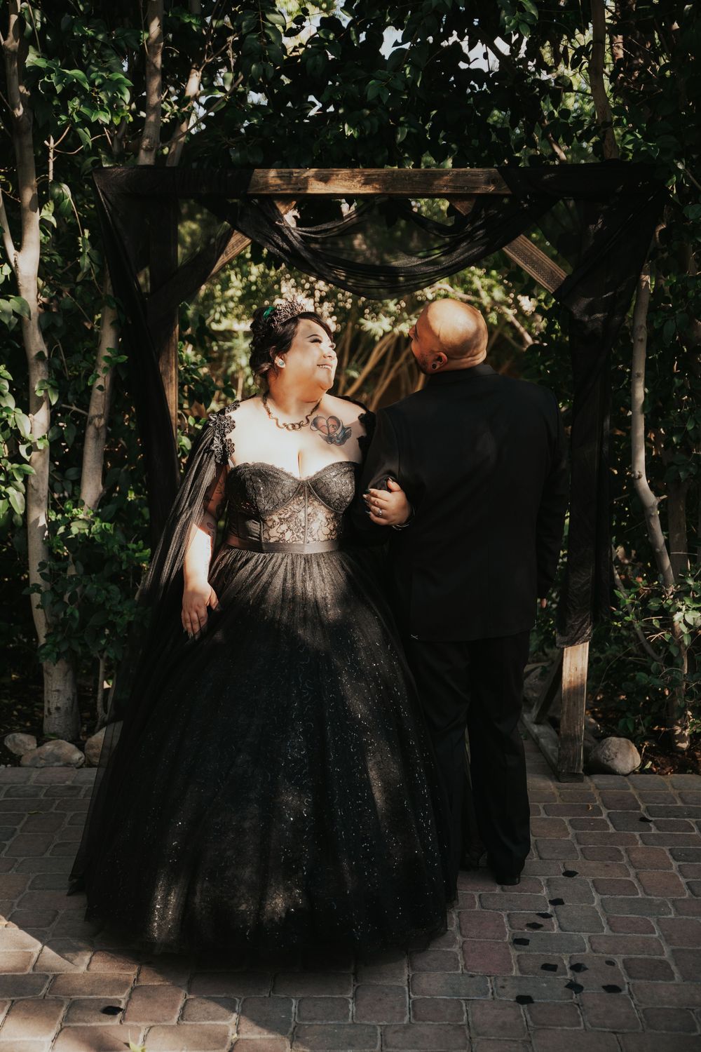A couple in formal attire shares an intimate moment on a brick patio surrounded by lush greenery.