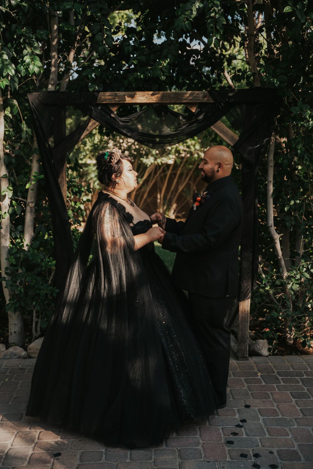 A couple in matching black formal attire dances together on a brick patio surrounded by lush greenery.
