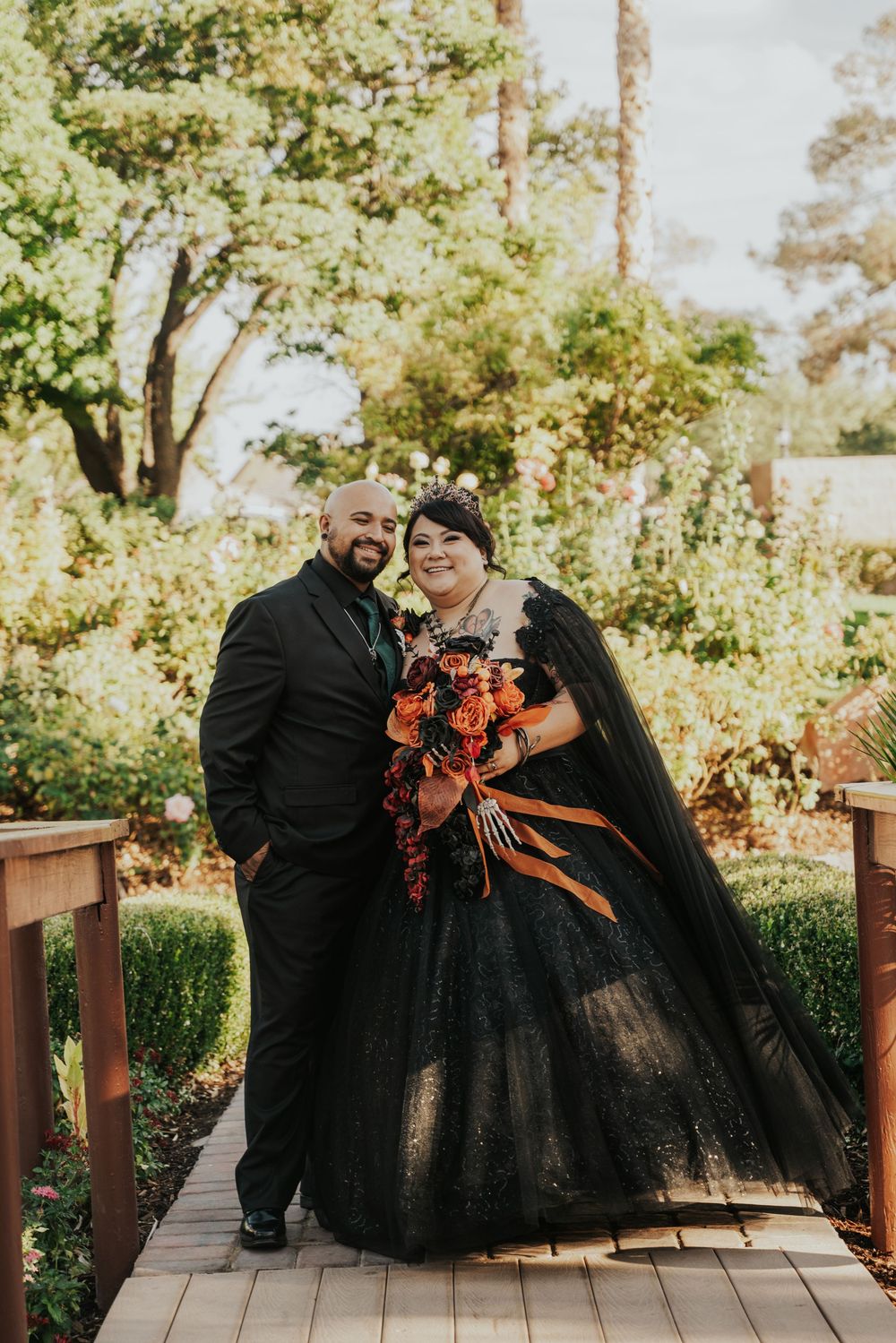 Couple in black formal wear shares intimate moments while posing on a wooden deck surrounded by greenery.