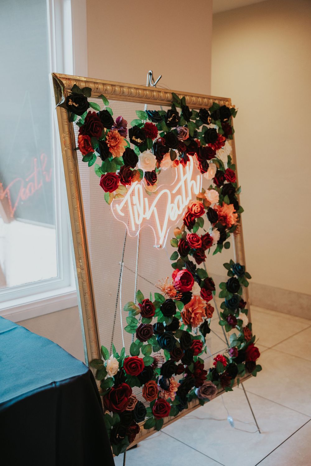A decorative frame with cascading red and pink roses and a glowing neon sign against a light colored wall.