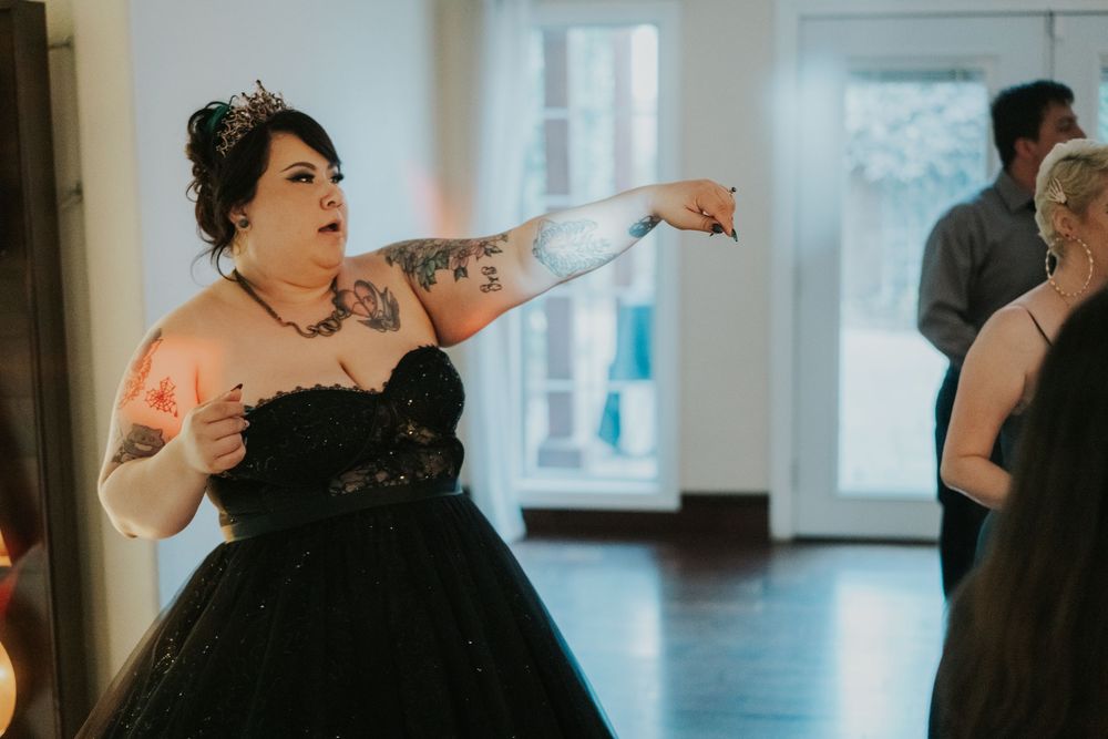 A person in a black formal dress gestures enthusiastically while dancing at an indoor event.