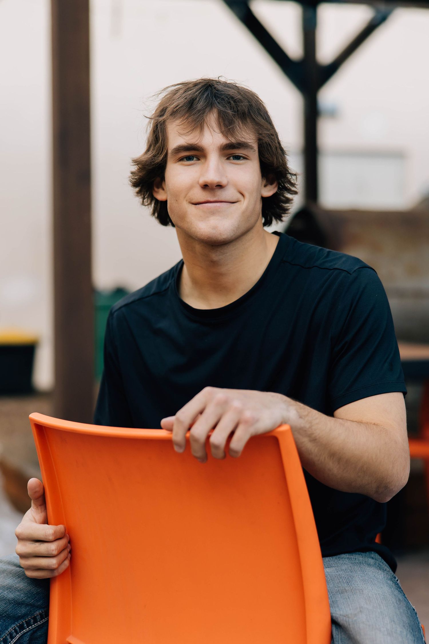 Smiling young man in black t-shirt sitting behind orange chair outdoors.