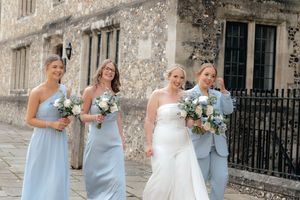 Wedding party in light blue dresses and white gown pose and laugh together outside a stone church building.