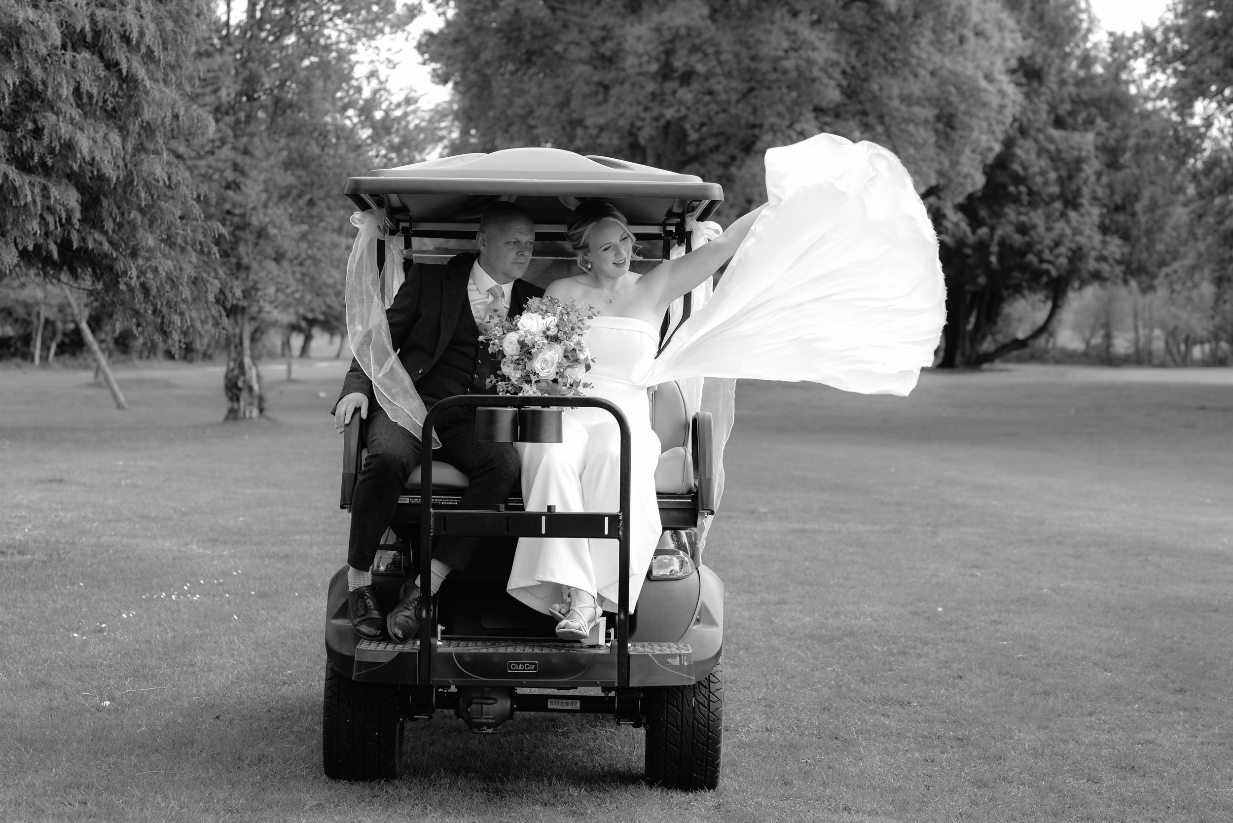 Golf cart drives across a grassy course during a sunny day with trees in the background.