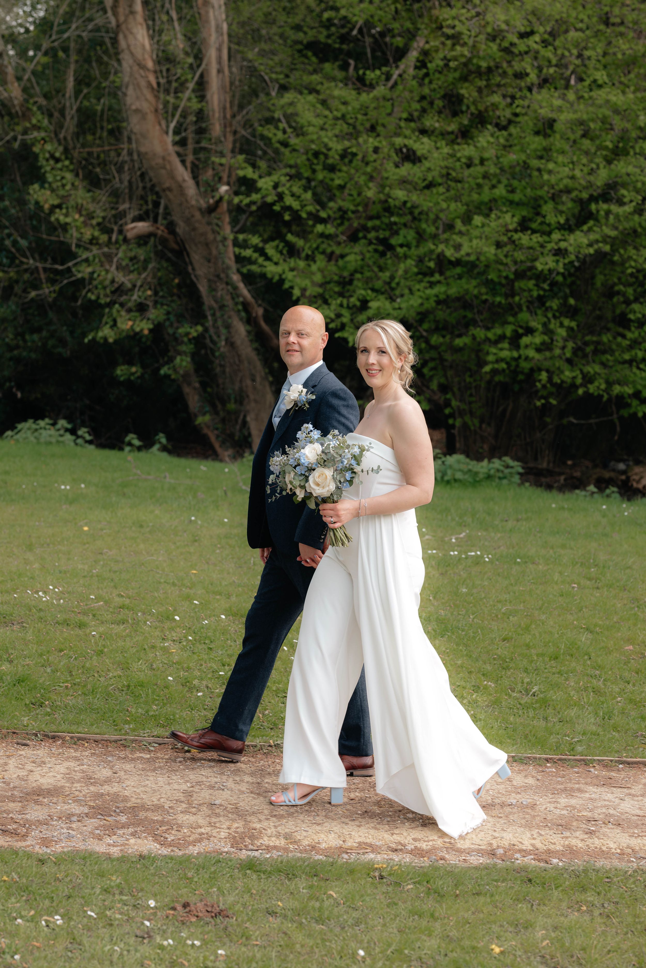 A couple poses on a dirt path during their wedding ceremony with green trees and grass in the background.