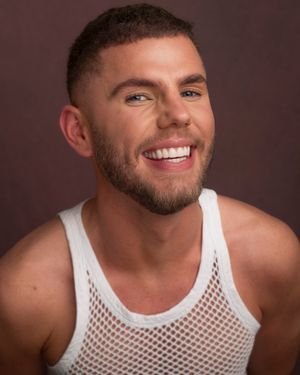 A cheerful person in a white mesh tank top smiles warmly against a dark brown studio backdrop.