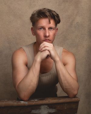 Portrait in muted tones showing figure in sleeveless top with clasped hands resting on wooden surface against textured backdrop.